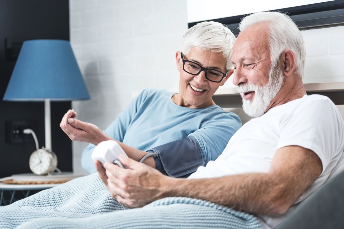 Senior couple in pajamas in a bed in a bedroom measure each other's blood pressure. Concept of home health check