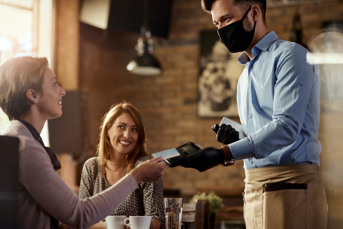 Young waiter wearing protective face mask while his guests are making contactless payment with credit card in a cafe.