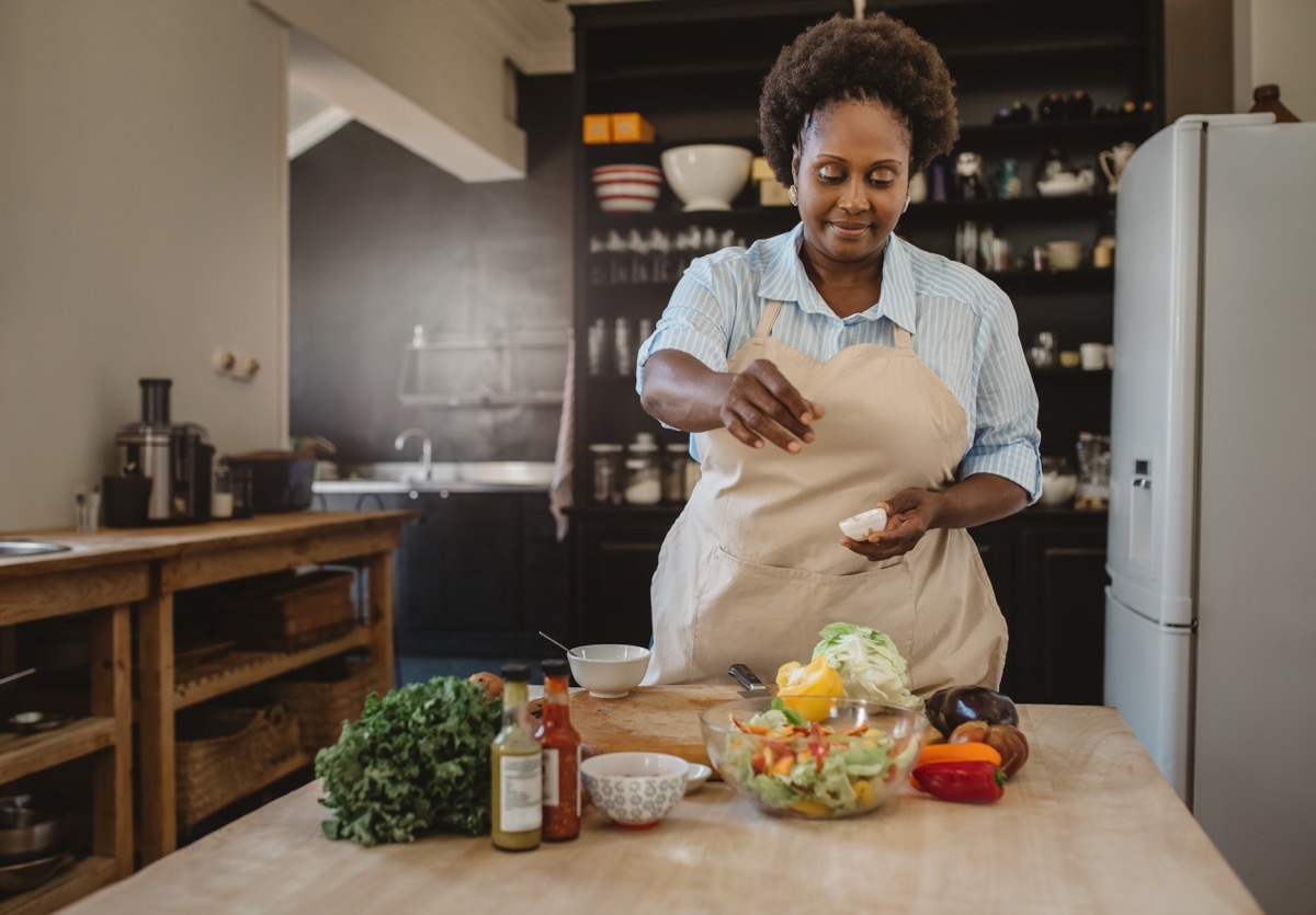 woman sprinkling seasoning over a delicious salad while standing at a counter in her kitchen at home