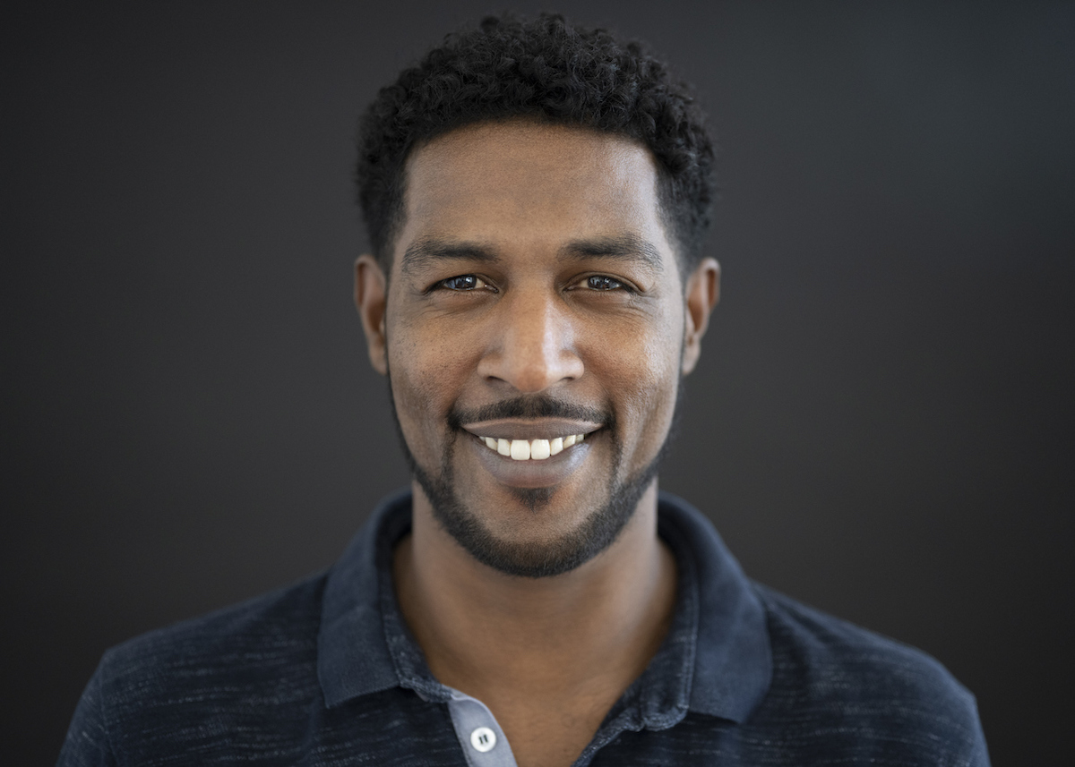 Front view of mature man with short hair and close-cropped beard and mustache wearing dark colored polo shirt and smiling at camera against black background.