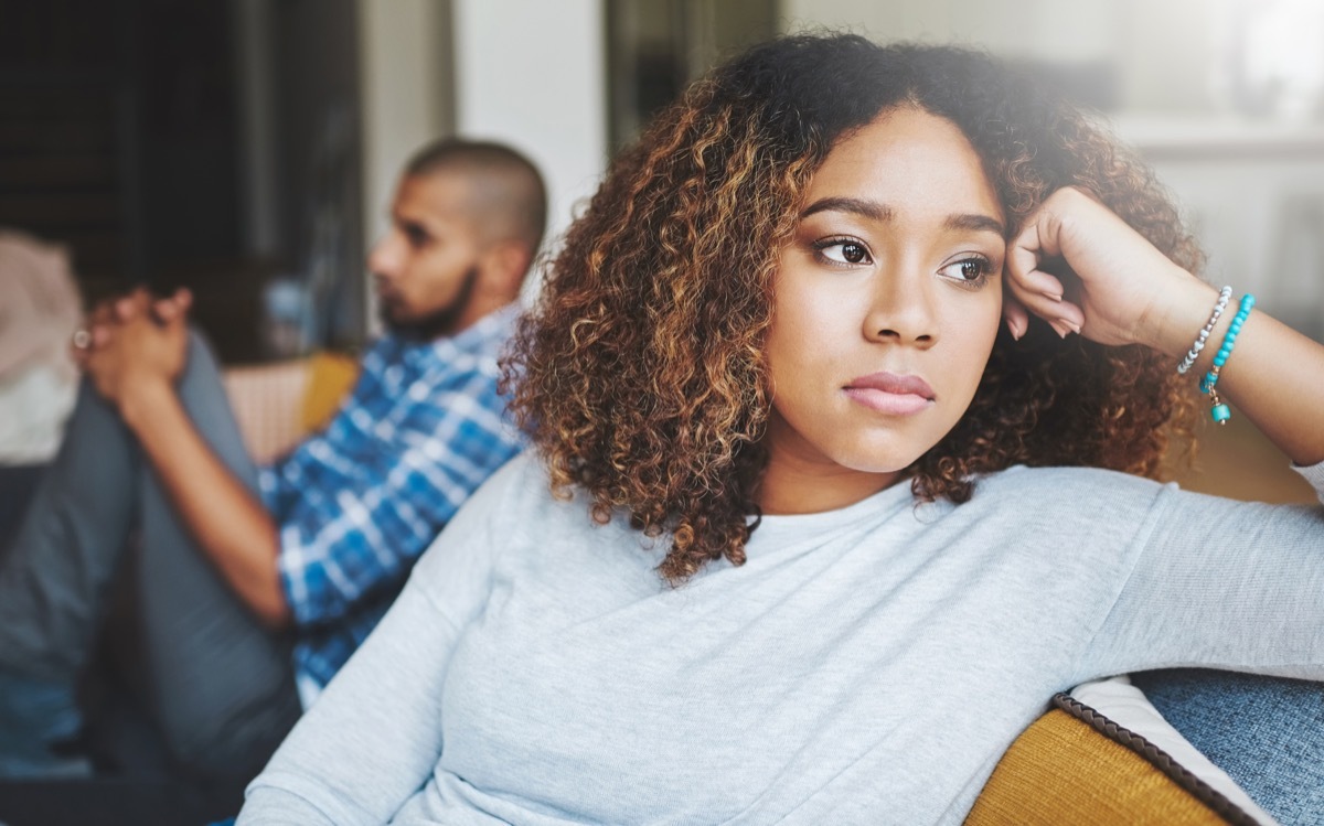 shot of a young woman looking upset after a fight with her husband in the background