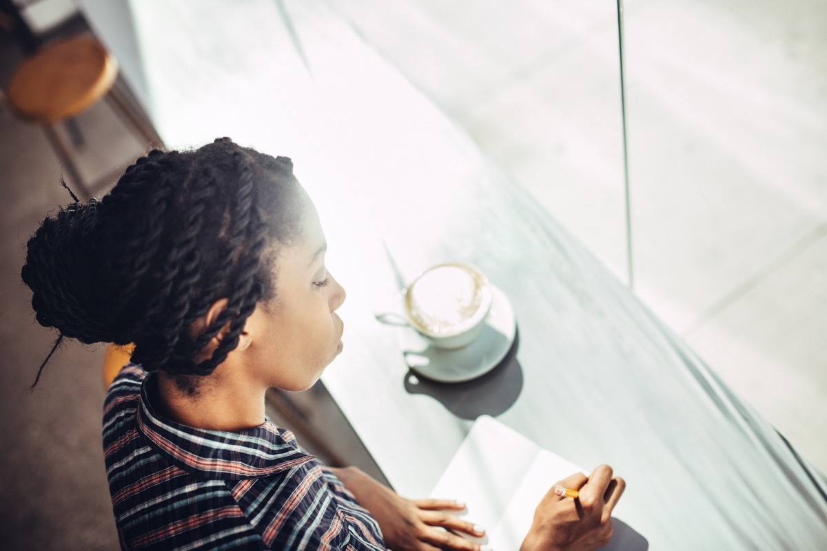 A beautiful young African American woman sits at the window counter of a coffee shop, enjoying a latte while writing ideas down in a small notepad journal or diary. She wears a casual button up shirt, with her hair up, and a content relaxed look on her face. Shot in downtown Los Angeles. Bright sunlight cascades in through the windows illuminating the pages of paper and hot drink. Horizontal with copy space.