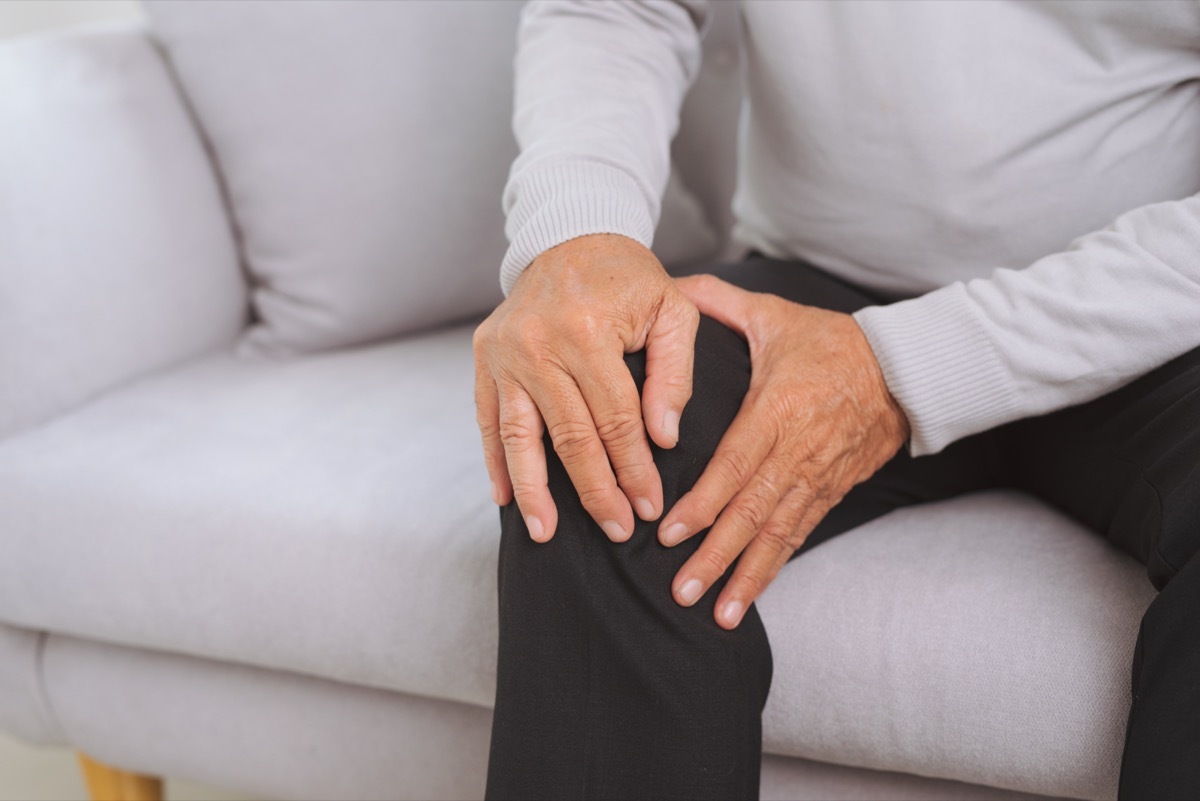 Senior man sitting on a sofa in the living room at home and touching his knee by the pain.