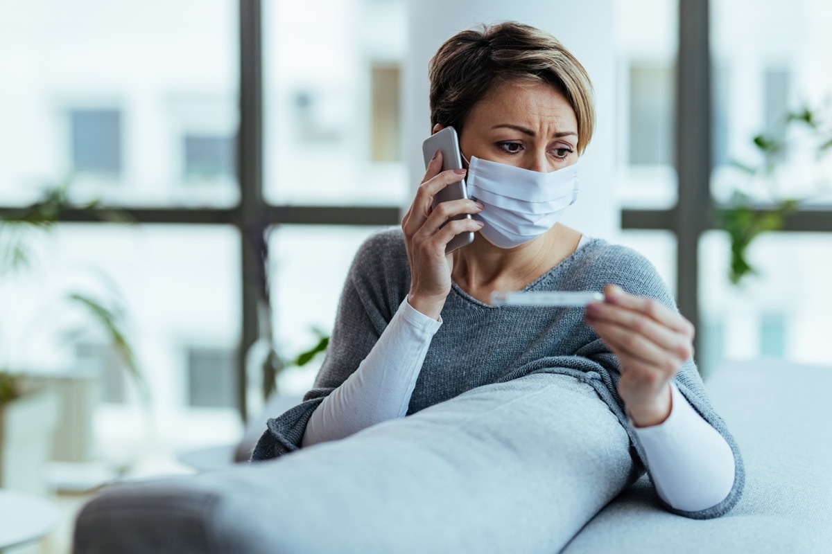 Distraught woman wearing face mask and measuring temperature while communicating on mobile phone at home.